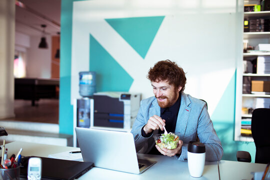 Businessman eating salad at laptop in modern office