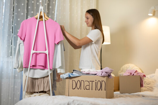 Teenage girl collecting and sorting her clothes for donation, placing garments into a cardboard box marked Donation.