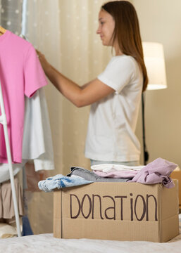 Teenage girl collecting and sorting her clothes for donation, placing garments into a cardboard box marked Donation.