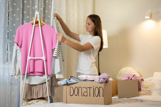 Teenage girl collecting and sorting her clothes for donation, placing garments into a cardboard box marked Donation.