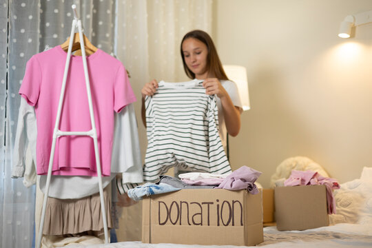 Teenage girl collecting and sorting her clothes for donation, placing garments into a cardboard box marked Donation.