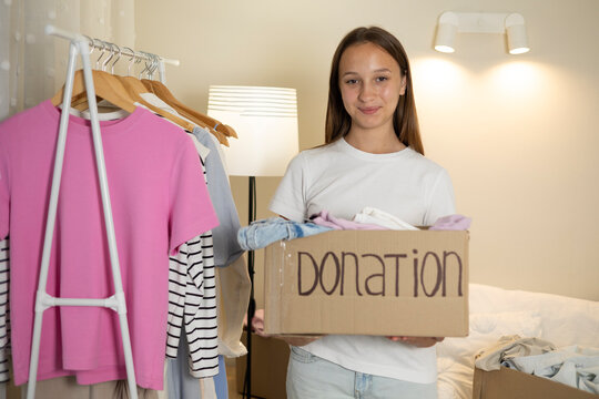 Teenage girl collecting and sorting her clothes for donation, placing garments into a cardboard box marked Donation.