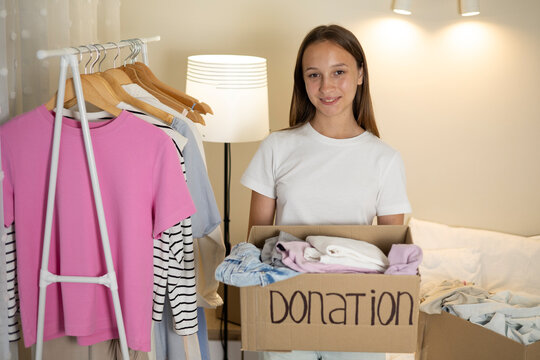 Teenage girl collecting and sorting her clothes for donation, placing garments into a cardboard box marked Donation.