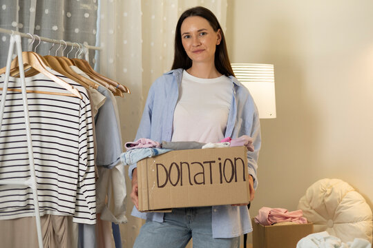 Young woman sorting and donating clothes at home, placing garments into cardboard donation boxes for charity and reuse.