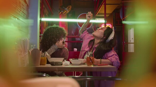 Young adult Asian women eating noodles with chopsticks and laughing in neon-lit cafe. Clip is perfect for food delivery apps, restaurant ads, and youth lifestyle marketing campaigns