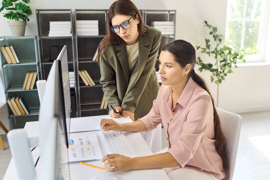 Women colleagues analyze analytics at computer workstation. Teamwork drives reporting as dashboards are reviewed in a light office. Teamwork collaboration and analytics concept for business.