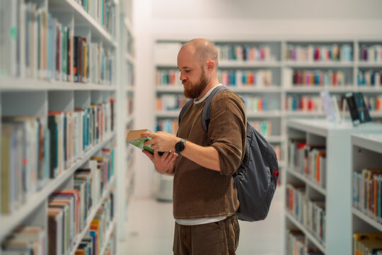 Man carrying an open book through a library aisle
