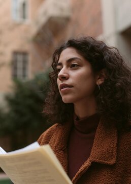 Portrait of a young woman with curly hair. she is standing outside, holding a book in her hands and looking off to the side with a thoughtful expression on her face.