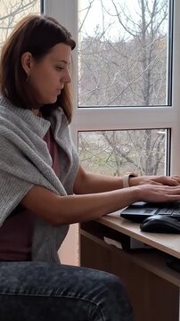 A woman works on a laptop while sitting by a window in a simple room