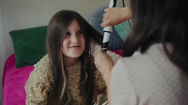 A mother gently curls her young daughter's hair with a curling iron at home, preparing for a festive celebration.
