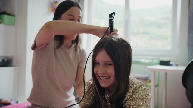A smiling mother straightens her laughing young daughter's hair with a flat iron in a bright room at home.