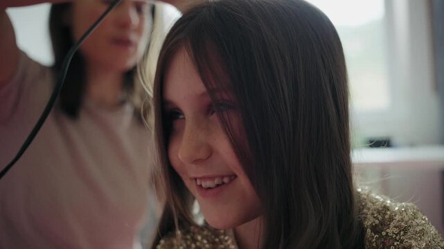 Mother gently styling the long hair of her smiling young daughter dressed in a sparkly sequin dress for a festive occasion at home.