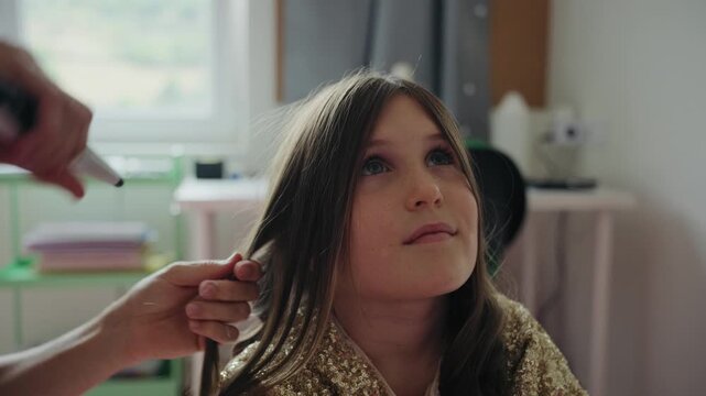 A mother styles and curls her young daughter's hair with a curling iron at home, preparing for a special occasion or party.