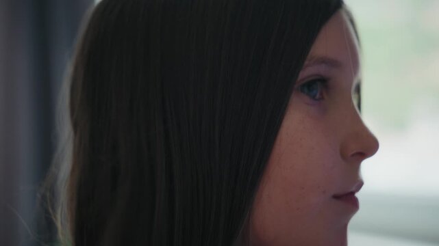 Close-up side view of a young girl sitting calmly while someone brushes her long dark hair with a wooden hairbrush at home.