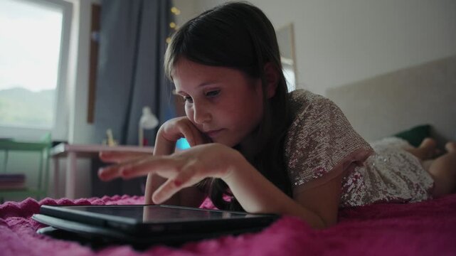 A young girl lies on a bed in her room while browsing and playing on a digital tablet touchscreen.