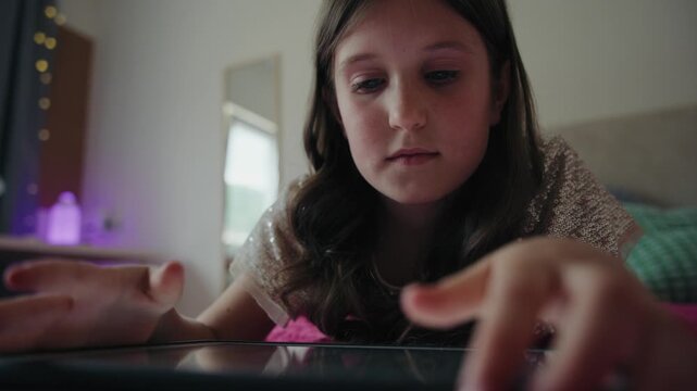 A focused young girl lies on a bed in her bedroom while browsing and playing on a digital tablet screen.