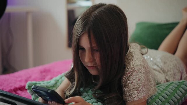 A young girl lies on a bed with pillows, focused on using a smartphone in a cozy bedroom.