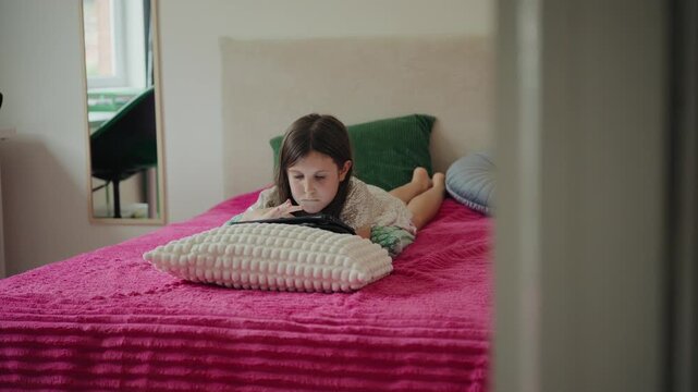 A focused little girl lies on a bed at home, browsing content on a digital tablet.