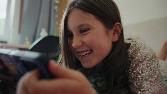 A cheerful young girl lies on her bed at home, watching funny videos on her smartphone and laughing happily.