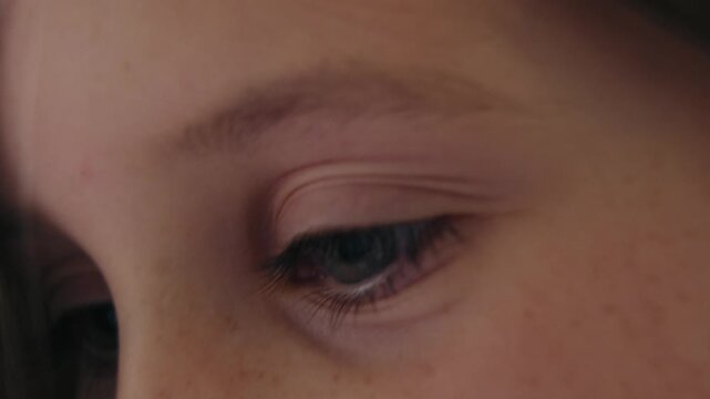 Macro close-up shot of a young Caucasian girl's blue eye, eyelashes, and freckled face as she looks down and blinks thoughtfully.