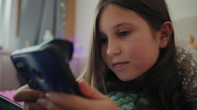A young girl lies comfortably in her bedroom while looking at her smartphone, smiling, texting, and watching videos on the screen.