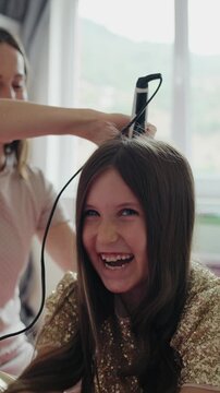 Vertical video of a smiling mother styling her happy daughter's hair with a flat iron at home.