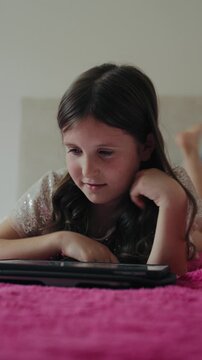 Vertical video of a focused little girl lying on a pink bed while tapping and browsing on a digital tablet at home.