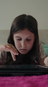 Vertical video of a focused young girl lying down on a bed while using a digital tablet at home.