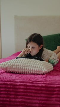 Vertical video of a young girl lying comfortably on a pink bed while using a digital tablet in her bedroom at home.