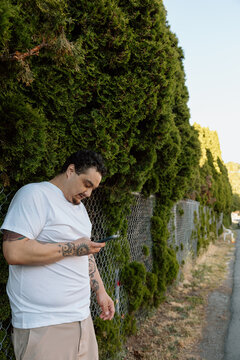 Candid man with tattoos checking phone by hedge and chain-link f