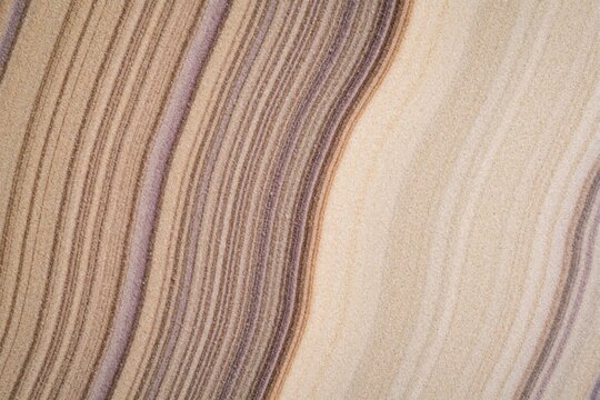 Close up view of natural sandstone texture. Wavy grain lines in beige tan, purple colors create abstract pattern. Sedimentary rock layers show geological erosion, mineral details. Smooth surface with