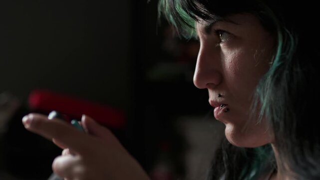 Profile view of a young woman with dyed hair and piercings intensely playing video games. Her face, lit by the screen, shows concentration and changing emotions