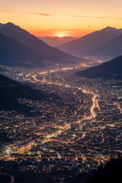 Aerial view of city lights spreading through valley at sunset. Mountains flank the illuminated urban sprawl. Warm orange sky fades into twilight above distant, glowing structures.
