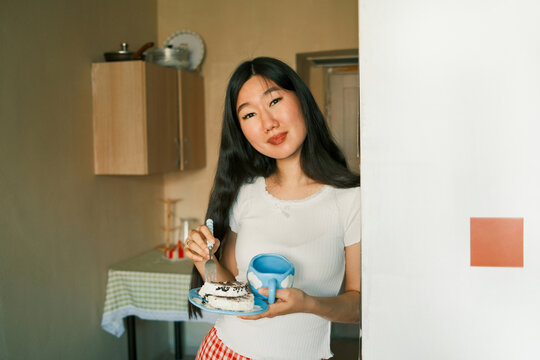 Young smiling asian woman eating cake and holding mug in the kitchen 