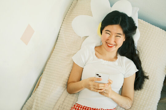 Overhead of happy young asian woman using smartphone lying in bed
