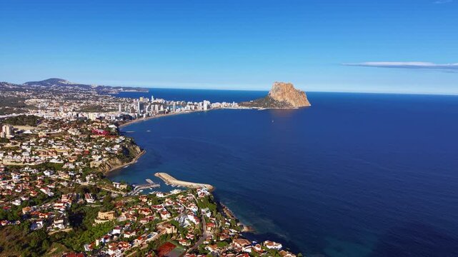 Panoramic view of Calpe, Spain, featuring the iconic Penon de Ifach rock, Mediterranean coastline, resort buildings, small marina, and residential neighborhoods under clear blue sky