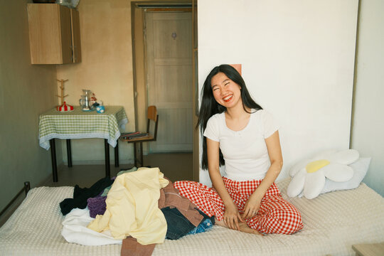 Happy young asian woman laughing, sitting on bed with clothes 