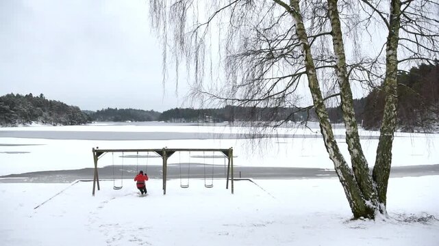 View of a person in a red jacket swinging on a wooden swing set at the snow covered Stora Delsjons badplats in Goteborg, Vastra Gotalands lan, Sweden.