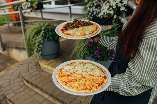 Restaurant worker carries two plated pizzas with assorted cheese, vegetable, corn, and sauce toppings through an outdoor dining area with flowers and greenery
