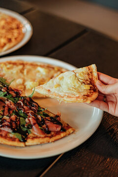 Hand lifts a hot pizza slice with melted cheese and sliced potato toppings from a mixed pizza on a plate, with fries and a mint drink blurred in the background