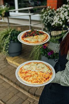 Restaurant worker carries two plated pizzas with assorted cheese, vegetable, corn, and sauce toppings through an outdoor dining area with flowers and greenery