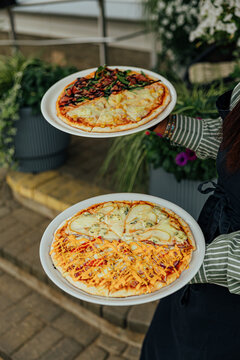 Restaurant worker carries two plated pizzas with assorted cheese, vegetable, corn, and sauce toppings through an outdoor dining area with flowers and greenery
