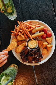 Overhead view of a large platter with fries, breaded bites, sausage pieces, dark toasted snacks, and creamy dipping sauce, with mint and lime drinks nearby