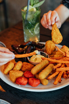 Hands reach for crispy fried snacks, fries, breaded bites, and sausage pieces arranged on a large plate with creamy dipping sauce at a dining table