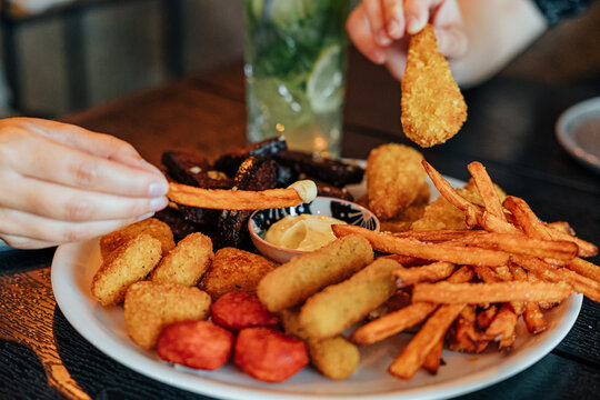 Hands reach for crispy fried snacks, fries, breaded bites, and sausage pieces arranged on a large plate with creamy dipping sauce at a dining table