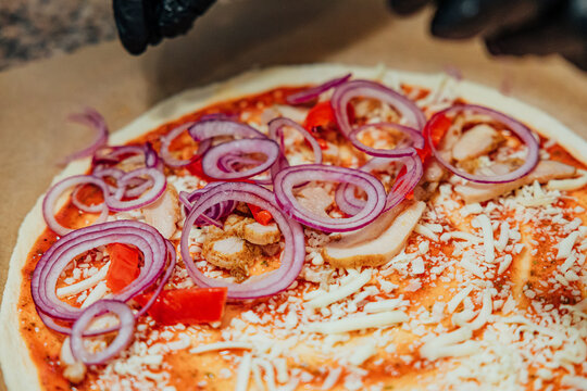 Black-gloved hands add red onion rings to an uncooked pizza topped with chicken, red pepper, shredded cheese, and tomato sauce