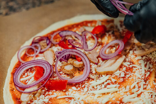Black-gloved hands add red onion rings to an uncooked pizza topped with chicken, red pepper, shredded cheese, and tomato sauce
