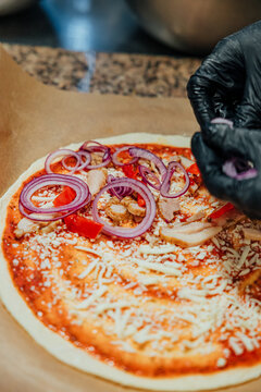 Black-gloved hands add red onion rings to an uncooked pizza topped with chicken, red pepper, shredded cheese, and tomato sauce
