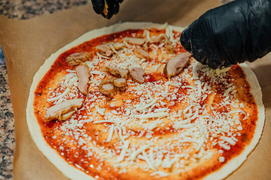 Black-gloved hands arrange sliced cooked chicken over uncooked pizza dough topped with tomato sauce and shredded cheese during kitchen preparation