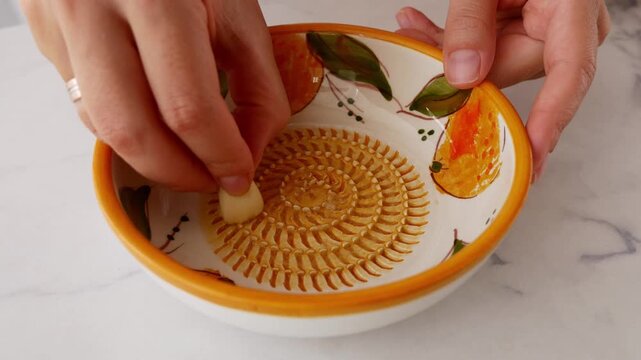 Woman grating fresh garlic on traditional Spanish ceramic grater plate. Close-up of authentic Mediterranean cooking and homemade food preparation